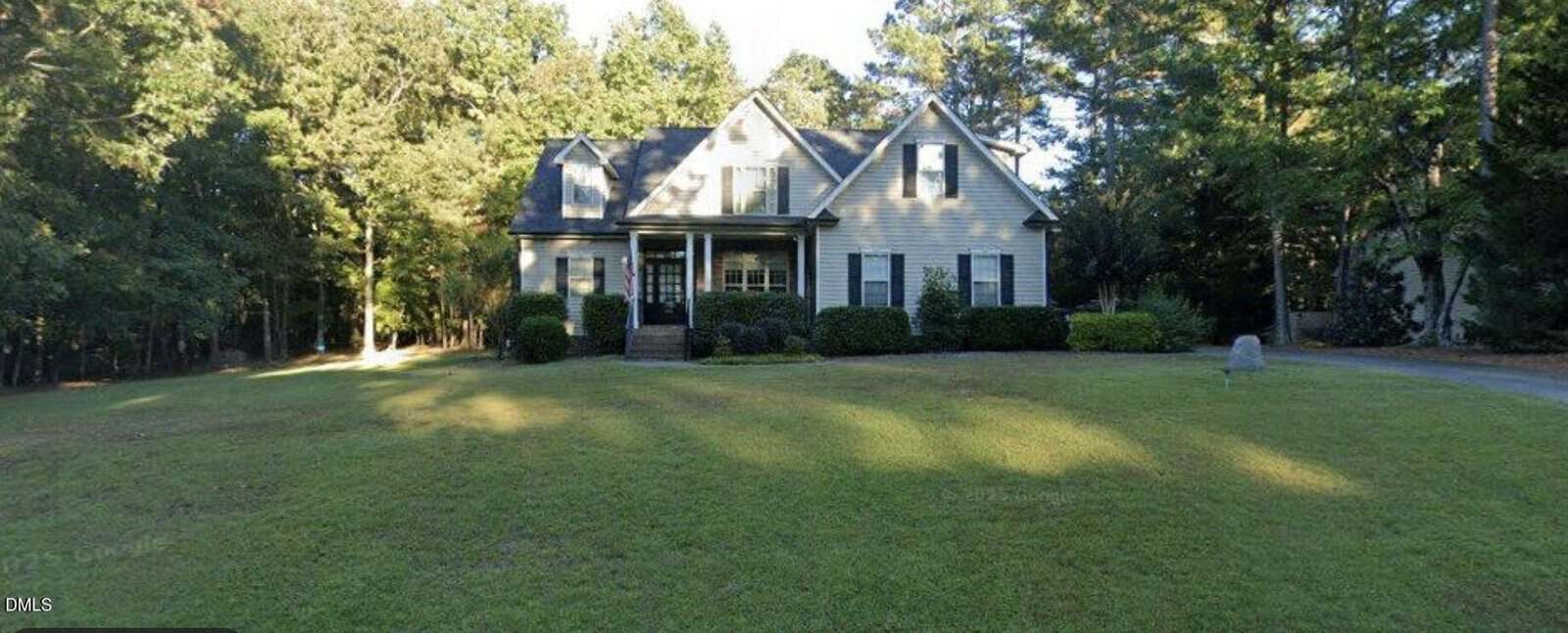 A two-story house with beige siding, dark shutters, and a covered front porch sits in the center of a large, neatly mowed lawn, surrounded by tall trees and greenery on a sunny day.