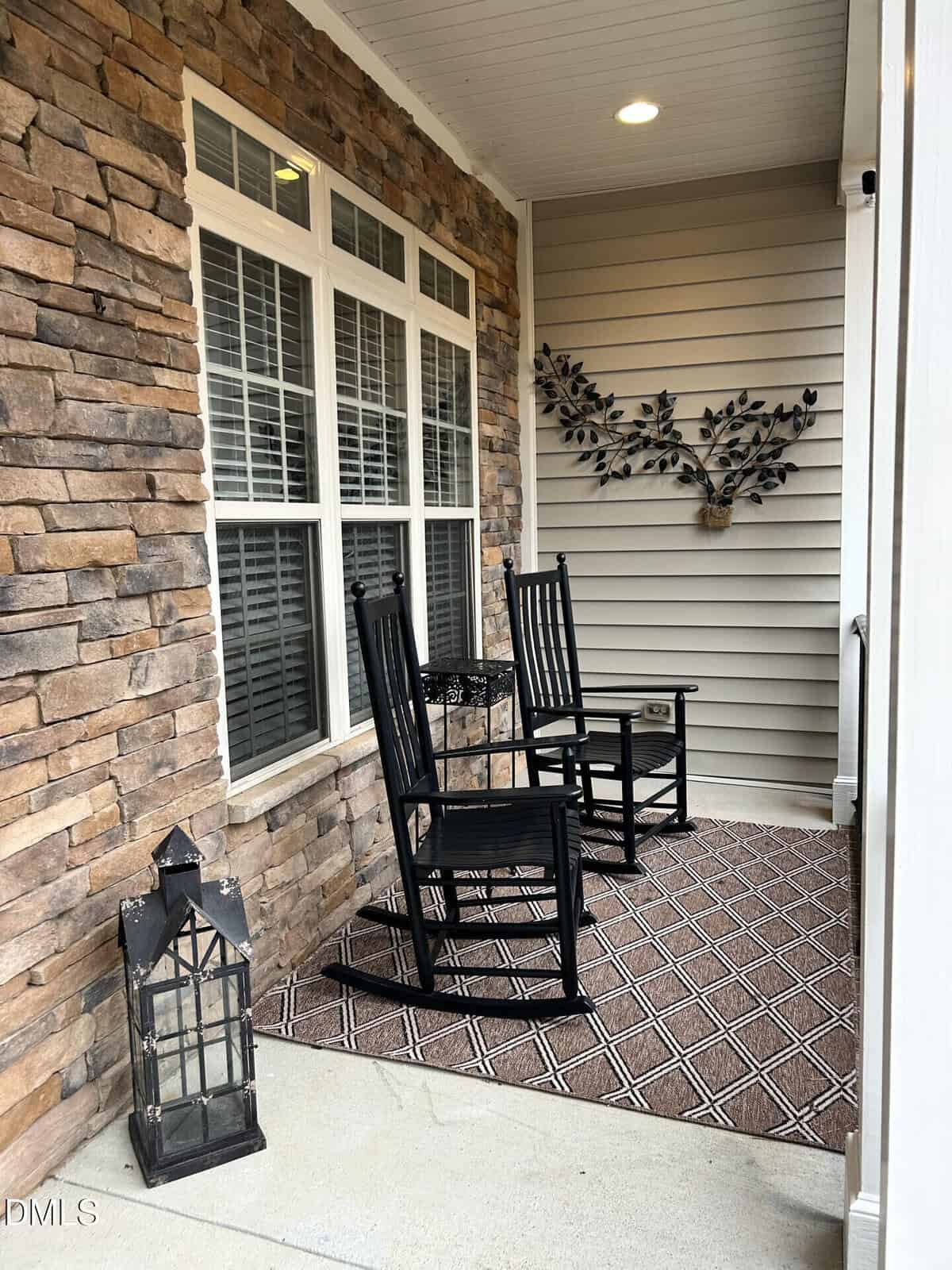A cozy porch with two black rocking chairs, a small table, and a patterned rug. A decorative branch wall art hangs on beige siding. Stone wall on the left; a large lantern sits near the entrance.