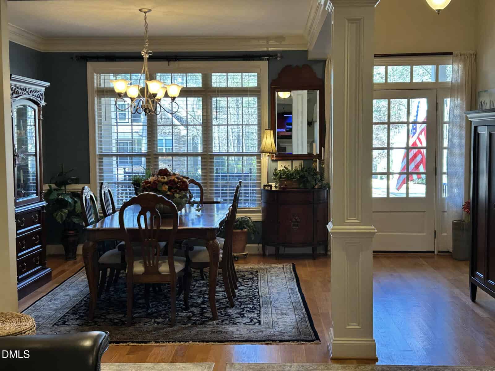 A dining room with a wooden table and six chairs sits on a rug near large windows. A chandelier hangs above, and a sideboard with a lamp and flowers is against the wall. An American flag is visible outside the door.
