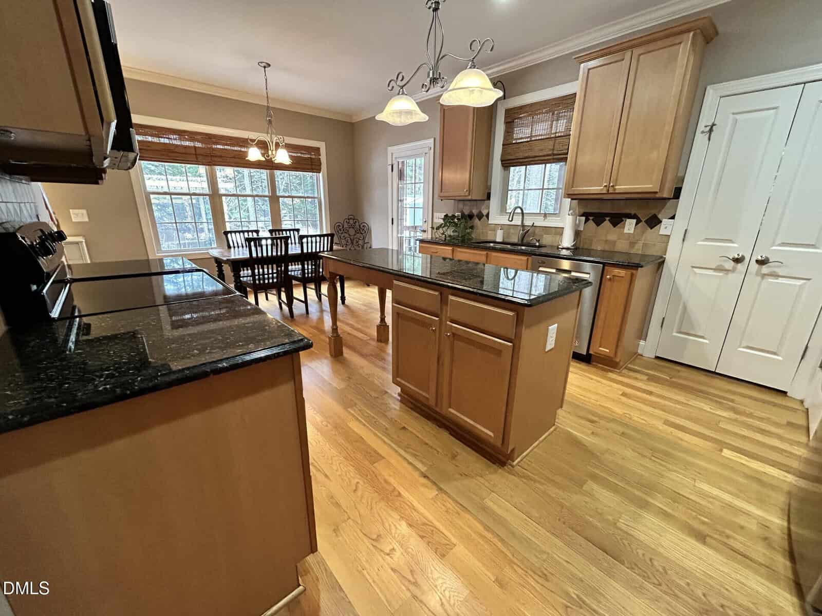 Spacious kitchen with wooden cabinets, black granite countertops, island, hardwood floors, and hanging lights. Adjacent dining area with table and chairs near large windows and a glass door.