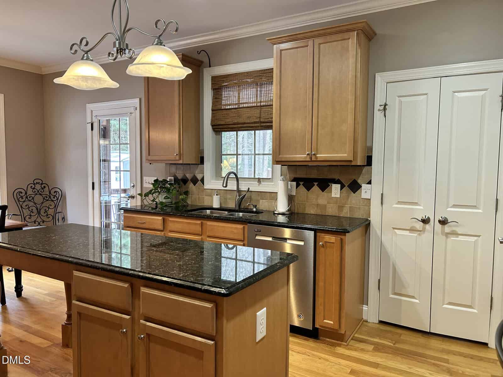 A kitchen with light wood cabinets, black granite countertops, a central island, stainless steel appliances, a window with a bamboo shade above the sink, and double pantry doors. Hardwood floors and pendant lights are visible.