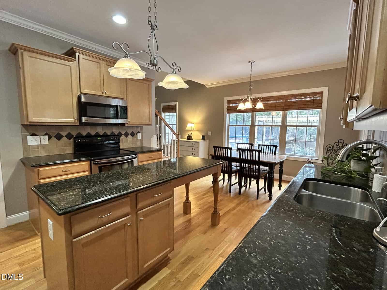 Spacious kitchen with light wood cabinets, black granite countertops, a center island, stainless steel appliances, and pendant lighting, opening to a dining area with large windows and a wooden table with black chairs.