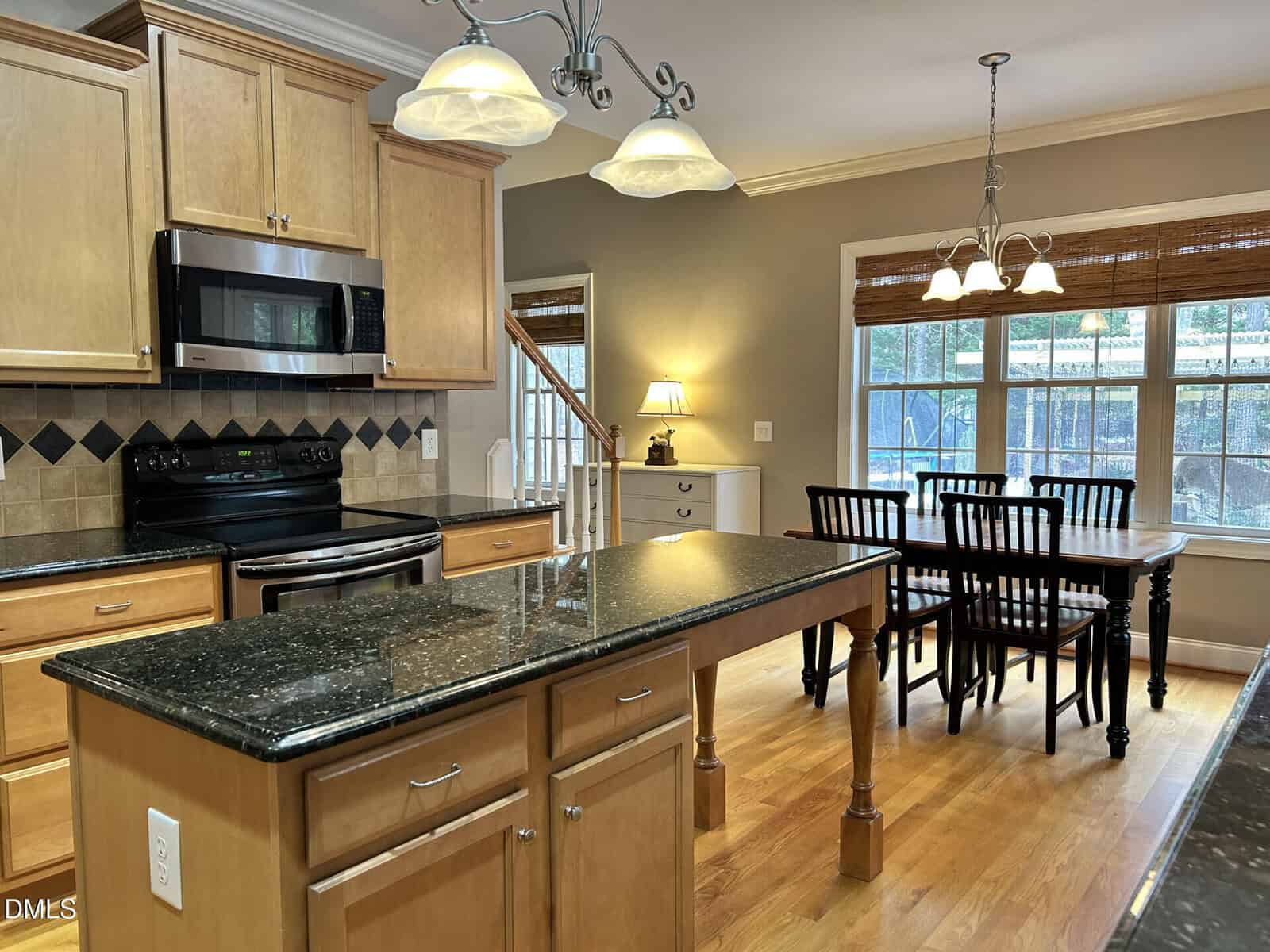 A spacious kitchen features light wood cabinets, black granite countertops, stainless steel appliances, a center island, pendant lights, and a dining area with a wooden table and six black chairs by large windows.