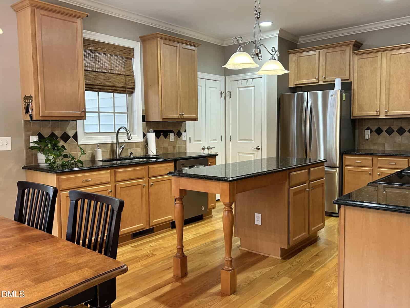 A modern kitchen with light wood cabinets, black countertops, an island with turned legs, stainless steel appliances, a double door pantry, and a small dining table with black chairs on a hardwood floor.
