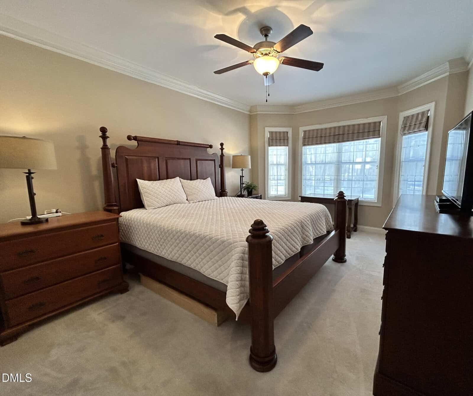 A bedroom with a large wooden bed, two matching nightstands with lamps, a ceiling fan, light-colored walls, carpeted floor, bay windows with blinds, and a dresser with a TV.