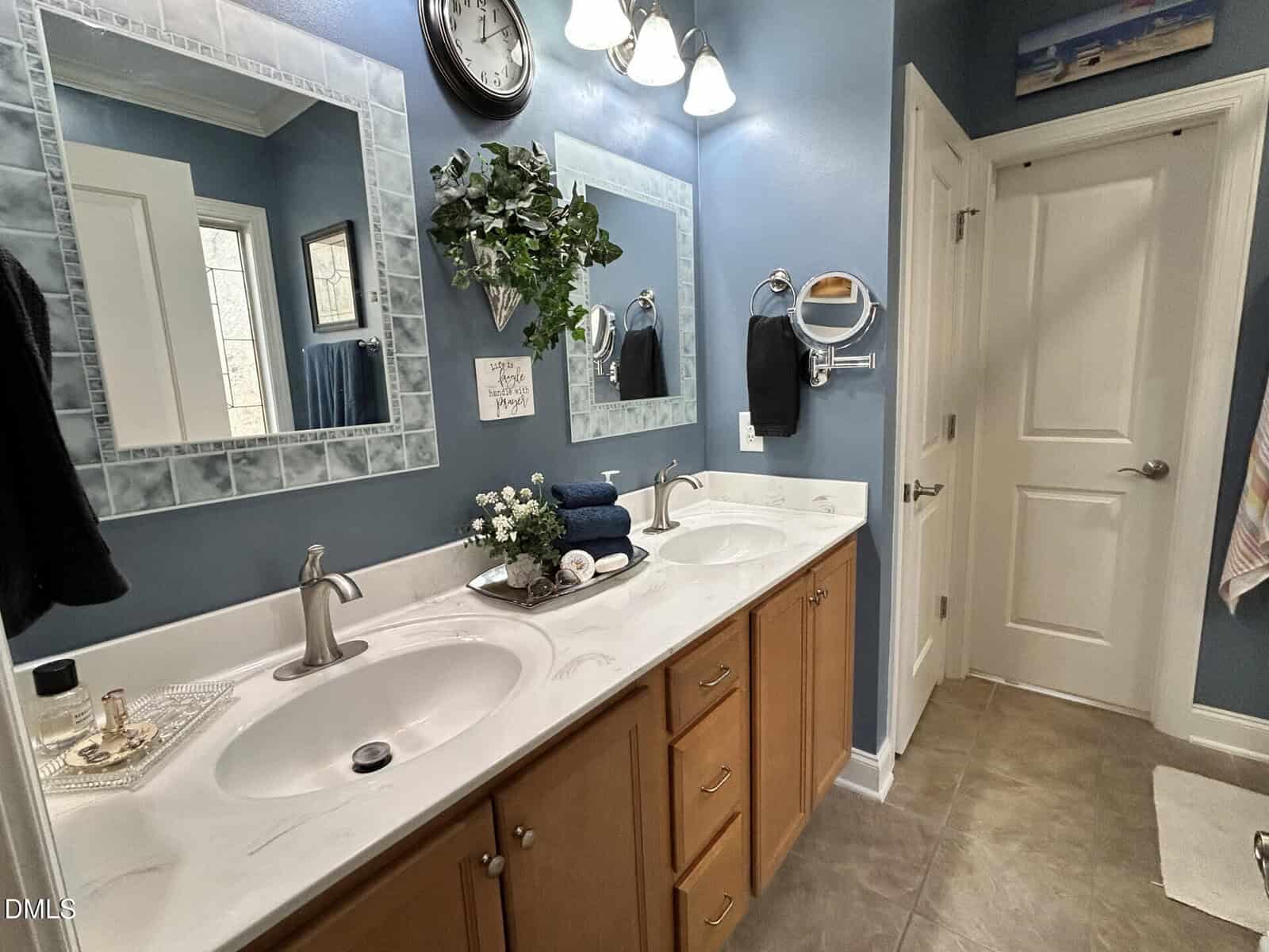 A blue bathroom with two sinks, a marble countertop, two mirrors, a wall clock, towels, and a plant above the sinks. Cabinets are below the counter, and a door is visible on the right.
