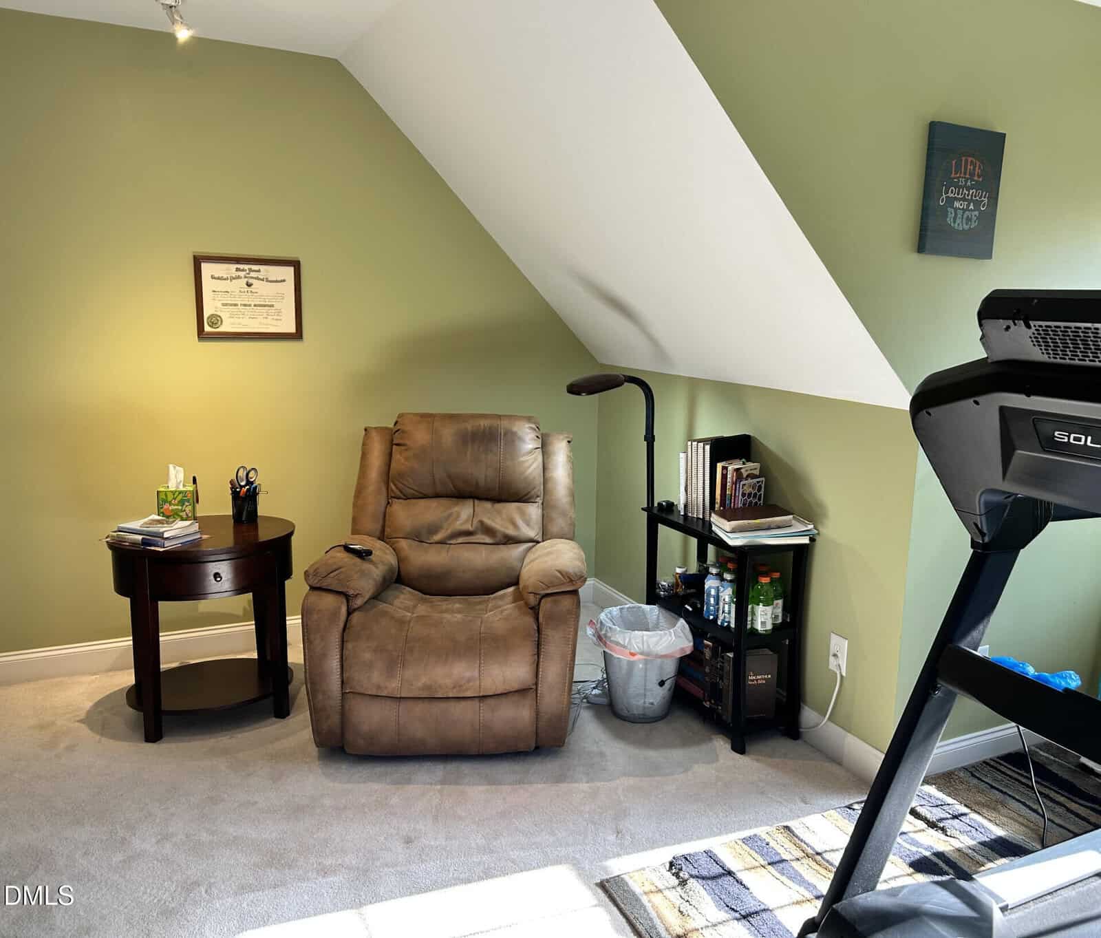A cozy home office with a tan recliner, side table, bookshelf, trash can, and treadmill under a sloped green wall. A framed certificate and inspirational art hang on the walls. Sunlight lights up the carpet.