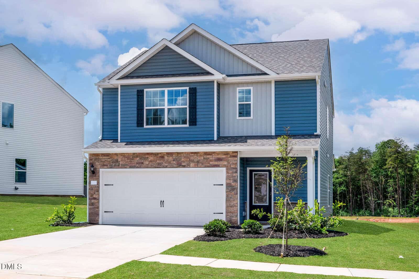 Two-story modern house with blue siding, brick accents, white trim, and a double garage. The front yard has a small tree, shrubs, and a concrete driveway leading to the garage. Blue sky and trees are visible in the background.