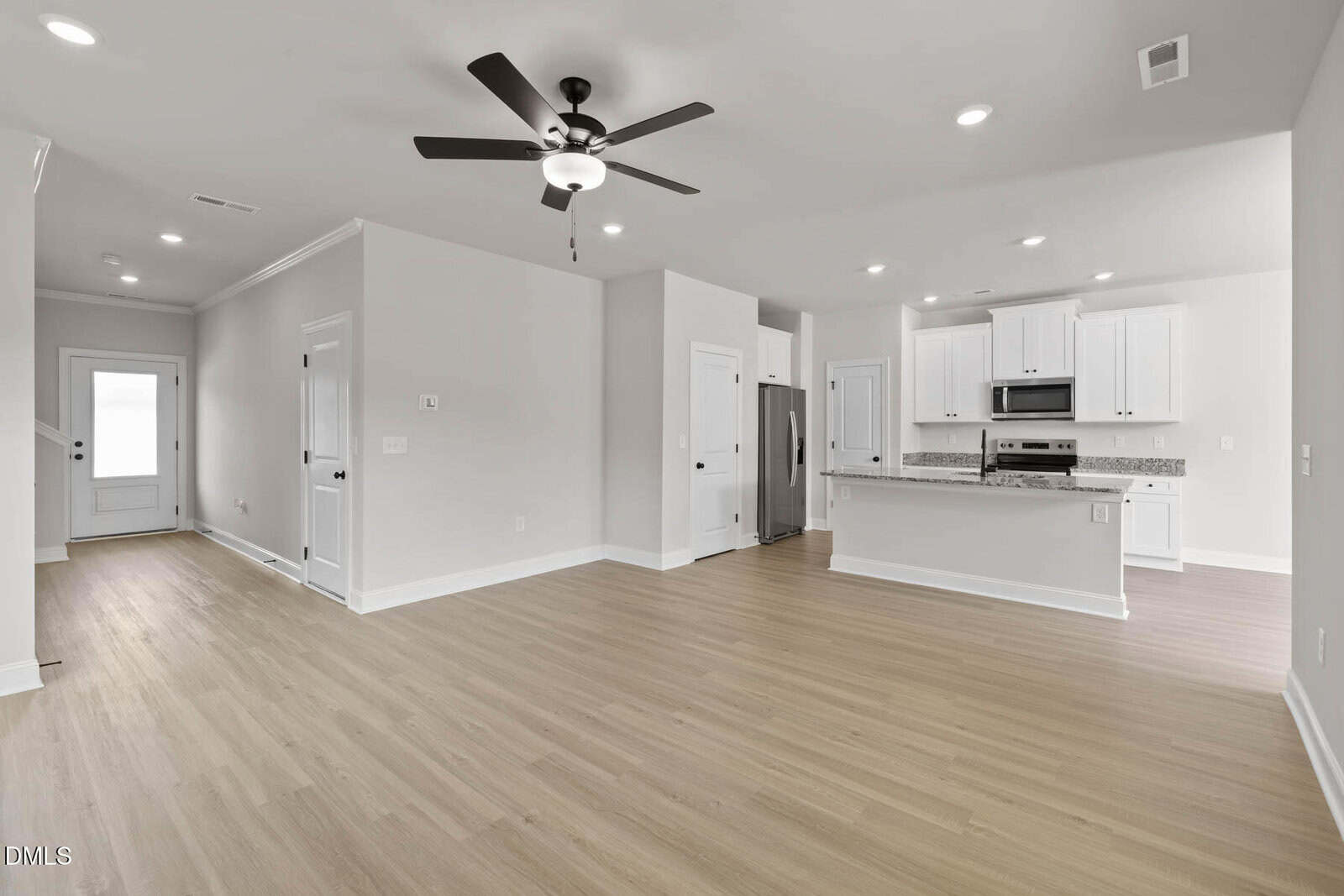 Bright, modern open-concept kitchen and living area with light wood flooring, white cabinets, granite countertops, ceiling fan, stainless steel appliances, and recessed lighting; a door and hallway are visible in the background.