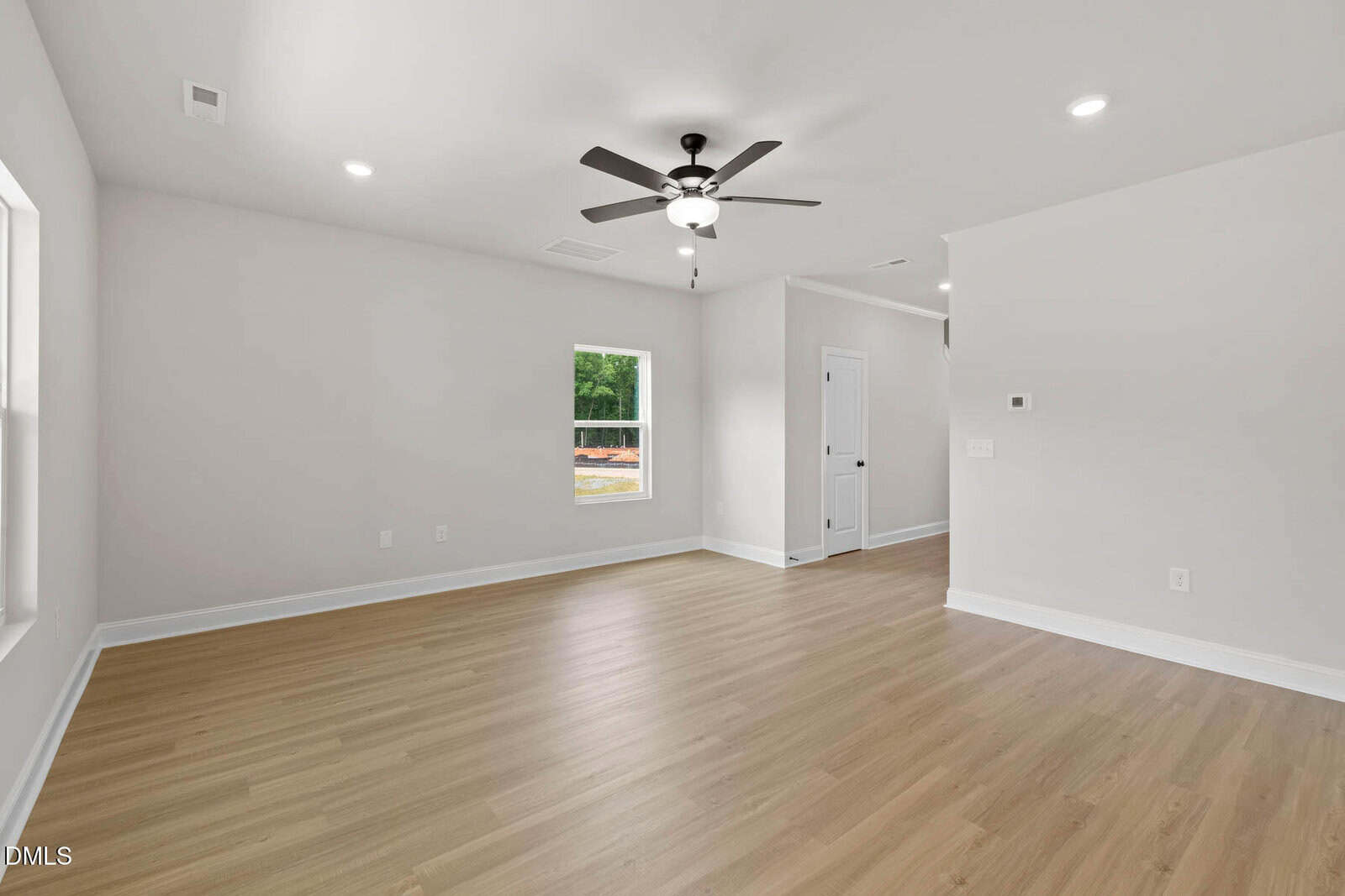 A bright, empty room with light wood flooring, white walls, recessed lighting, a ceiling fan with lights, and a window letting in natural light. A white door and hallway are visible in the background.
