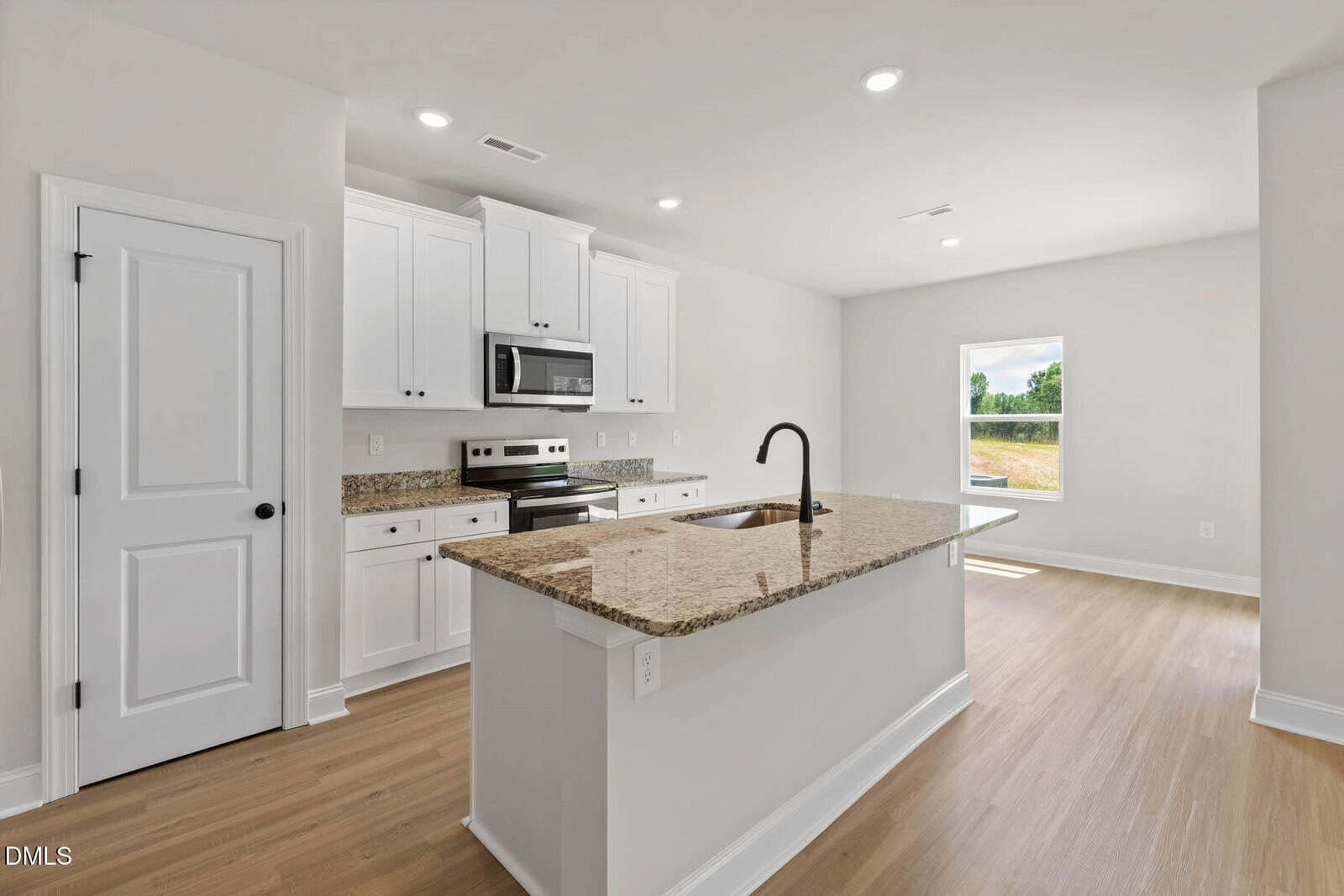 Bright, modern kitchen with white cabinets, stainless steel appliances, a built-in microwave, granite countertops, an island with a sink and faucet, and wood-look flooring. A window provides natural light.