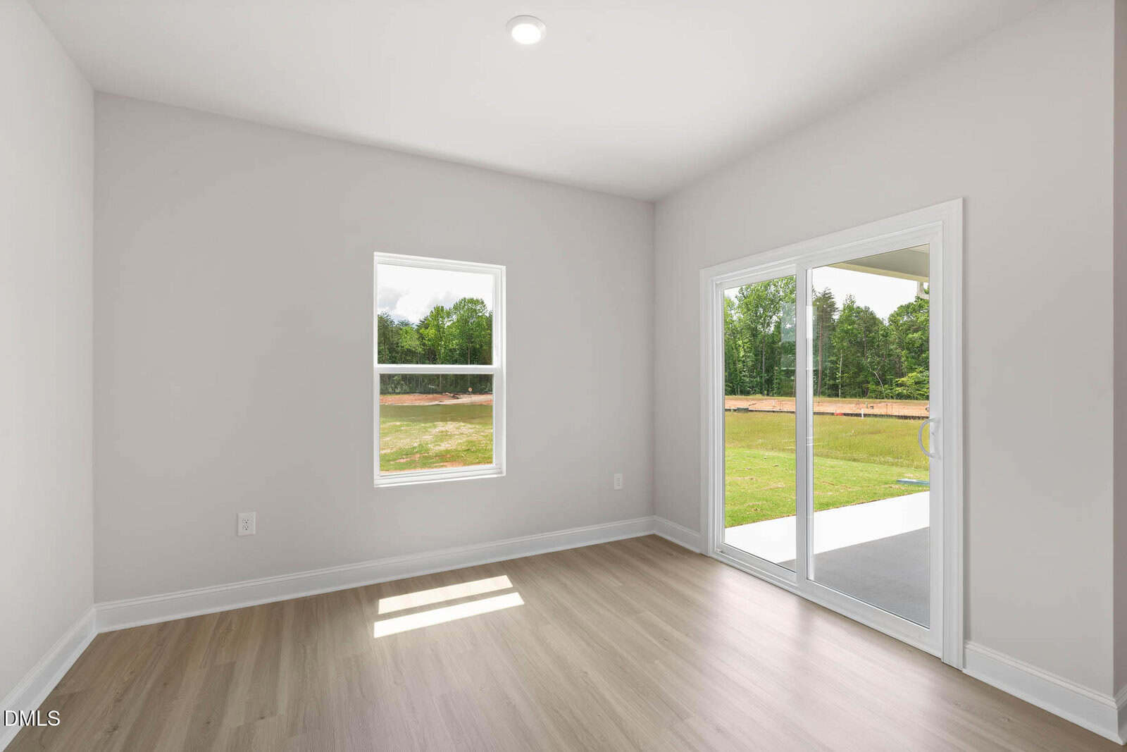 Bright empty room with light wood flooring, white walls, a single window, and large sliding glass doors opening to a patio with a green lawn and trees visible outside.