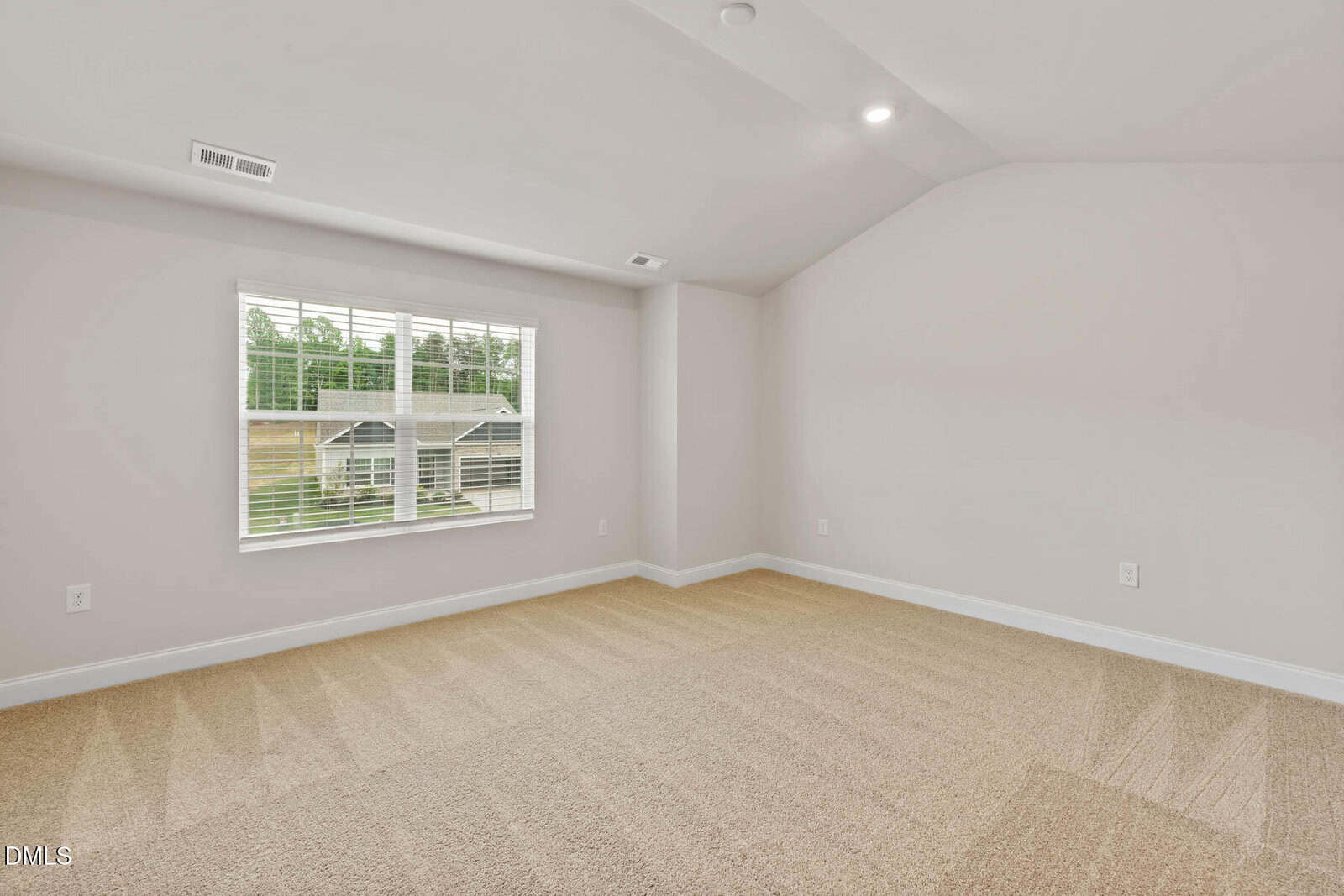A bright, empty room with beige carpet, light-colored walls, a large window with blinds letting in natural light, and a sloped ceiling with recessed lighting.