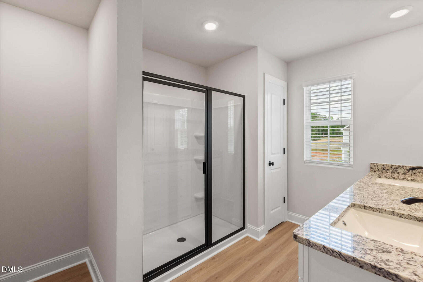 A bathroom with a glass-enclosed shower, a double-sink granite vanity, wood-style flooring, white walls, a small closet door, and a window with white blinds letting in natural light.