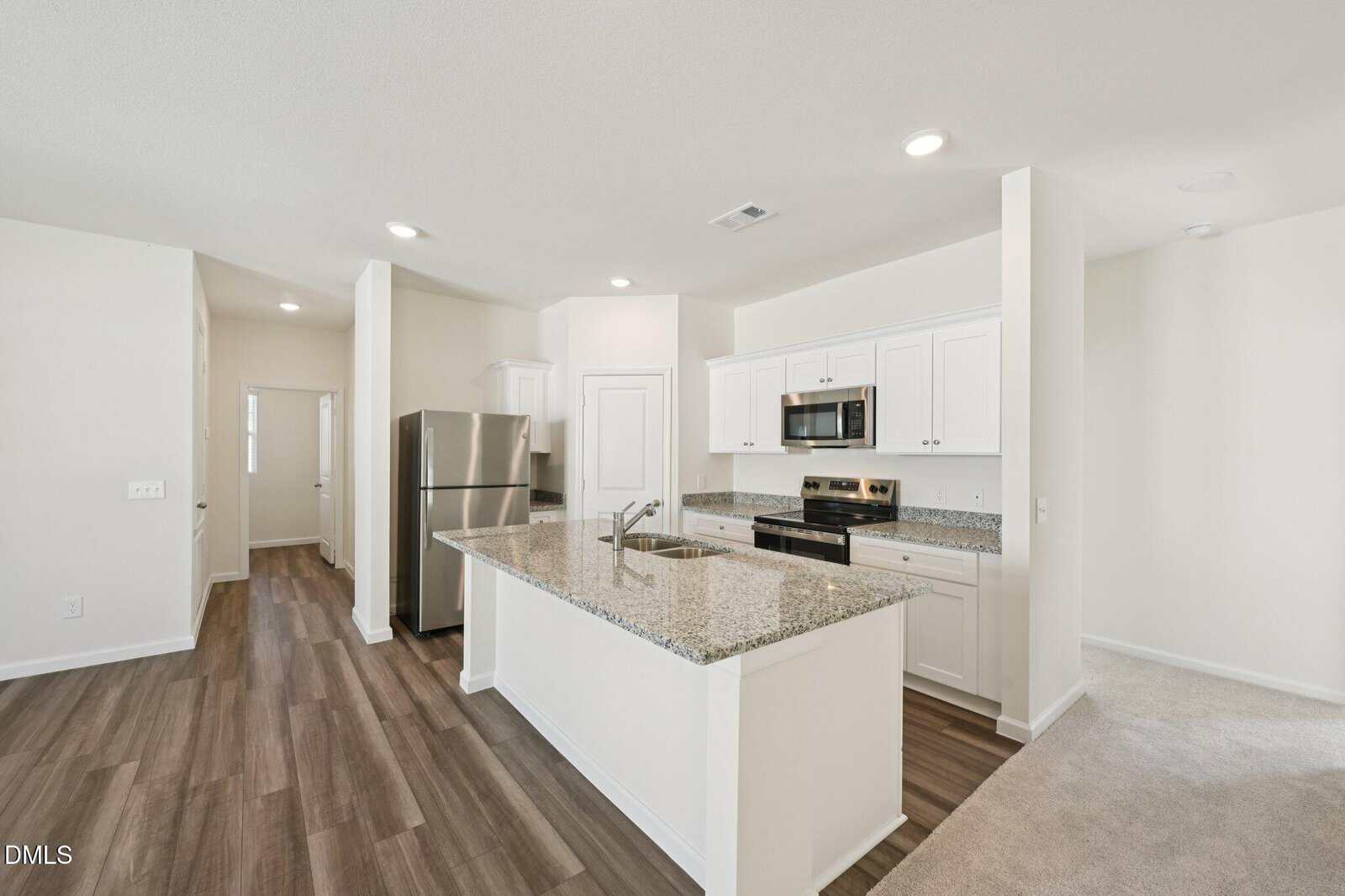 Modern kitchen with white cabinets, stainless steel appliances, granite countertops, and a center island. The floor is wood-style with adjacent carpeted area, and the space is brightly lit with recessed lighting.