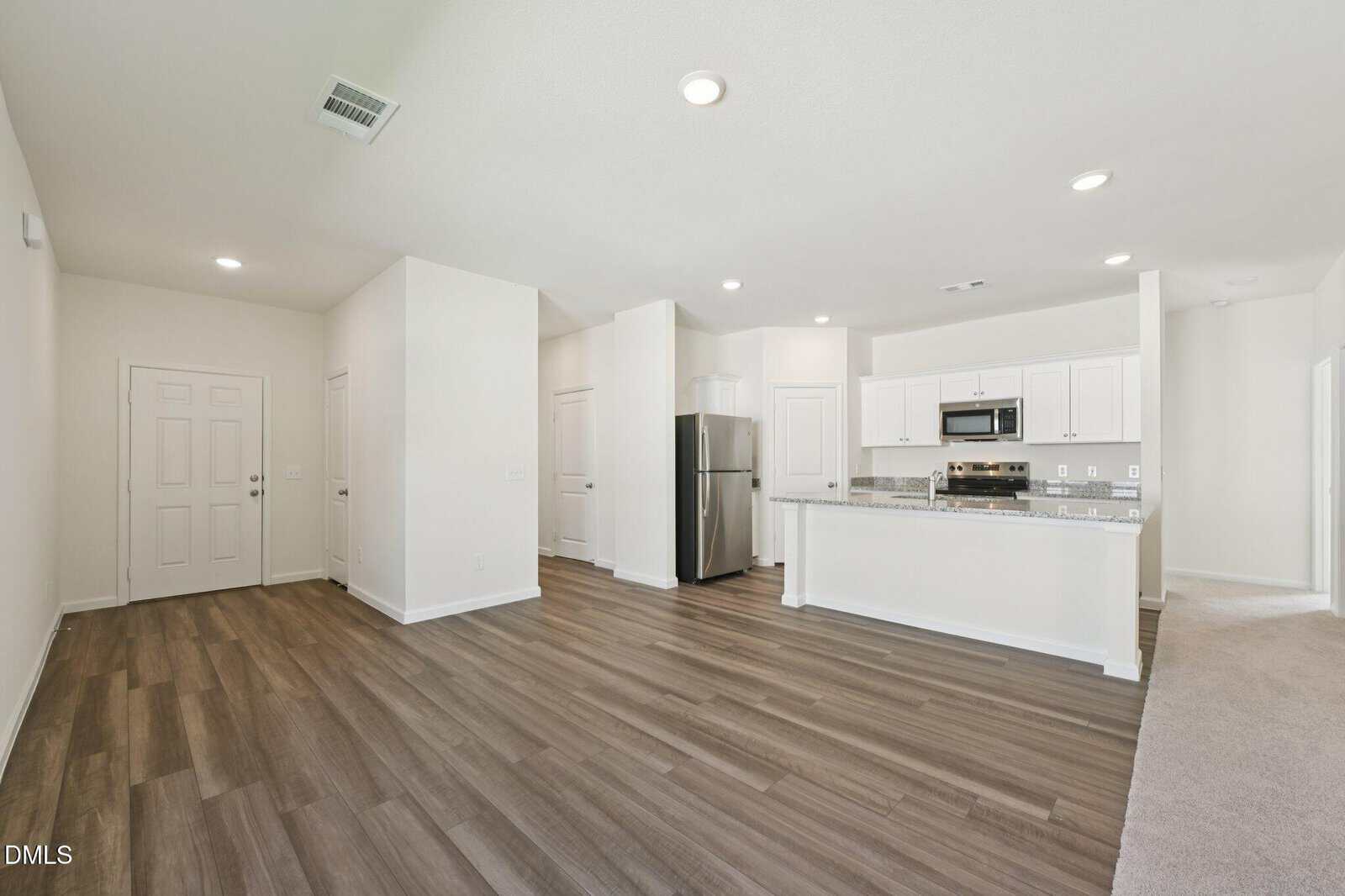Modern open-concept kitchen and living area with white walls, stainless steel appliances, white cabinetry, and wood-style flooring. The space is brightly lit with recessed lights and features a breakfast bar.