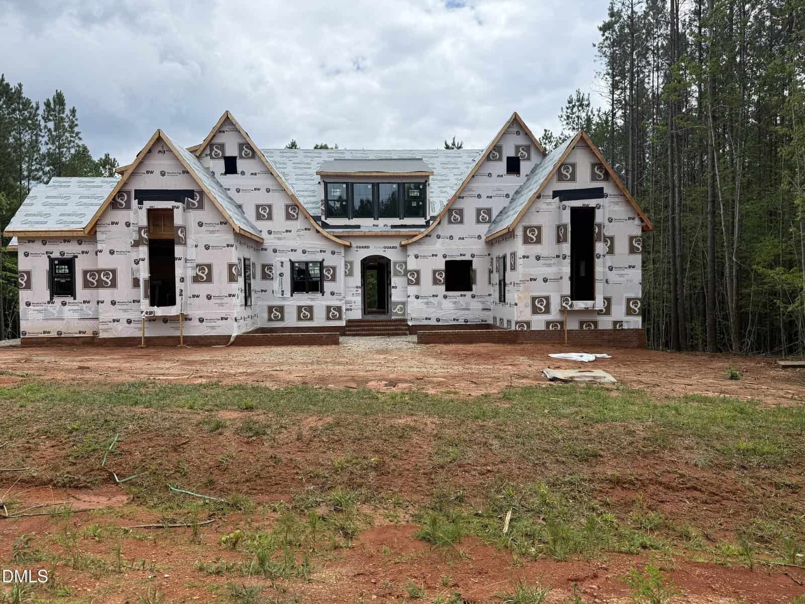 A large, two-story house under construction with white weatherproofing wrap and no exterior siding, set on a cleared, reddish dirt lot with trees in the background under a partly cloudy sky.