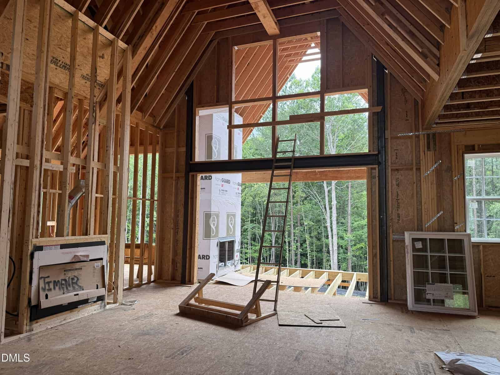 Interior of a house under construction, showing exposed wooden framing, a large window opening, a ladder, an unfinished fireplace, and construction materials scattered around. Trees are visible through the window openings.