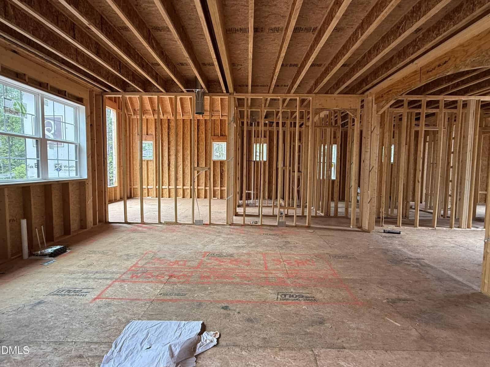 Interior view of a house under construction with exposed wooden framing, beams, and unfinished floors. Sunlight streams in through large windows on the left. Red markings are visible on the floor.