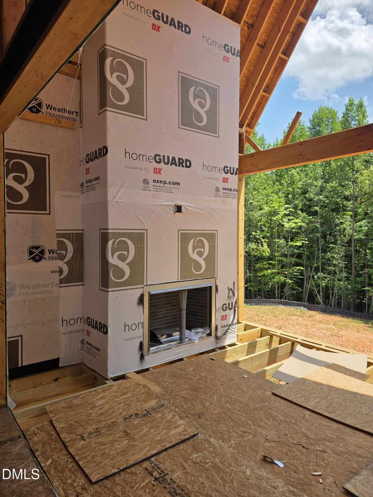 A partially constructed outdoor fireplace on a covered deck, surrounded by plywood flooring and exposed wooden beams, with trees visible in the background.
