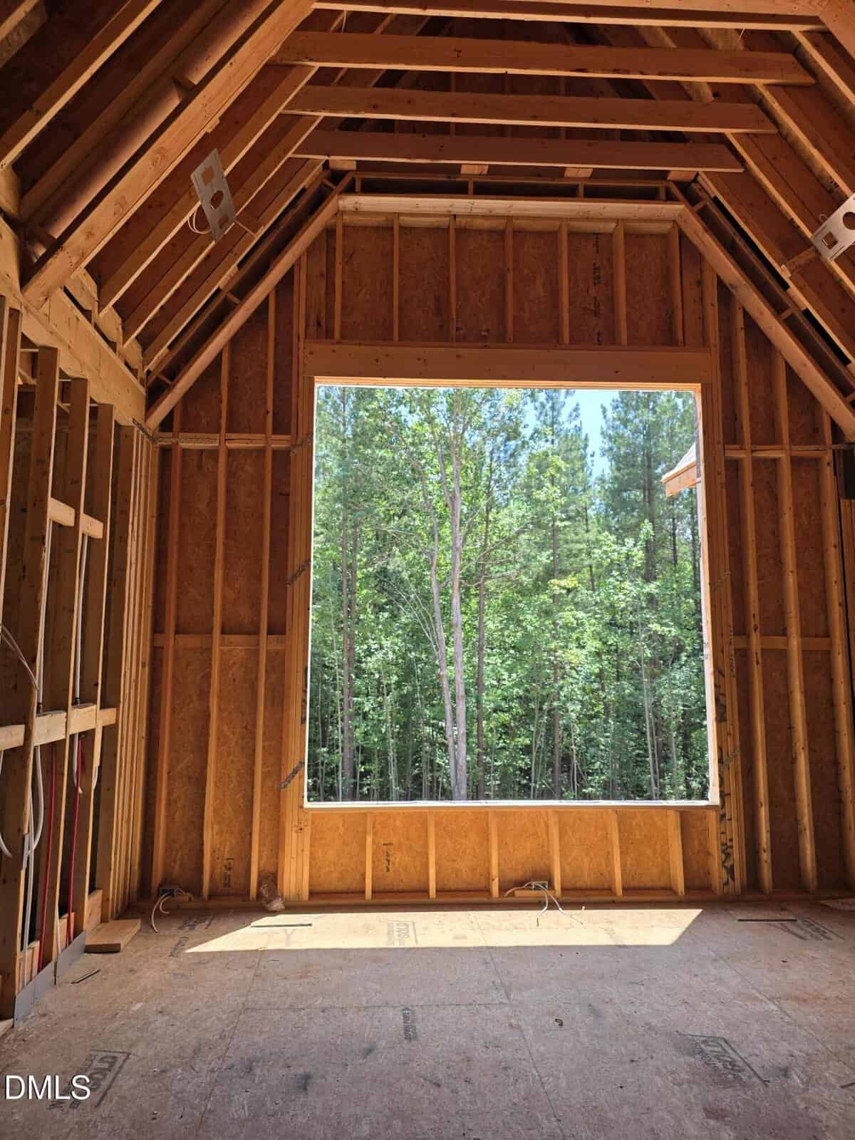 Room under construction with exposed wooden framing and a large square window opening. Through the window, tall green trees and a forested area are visible outside in bright daylight.