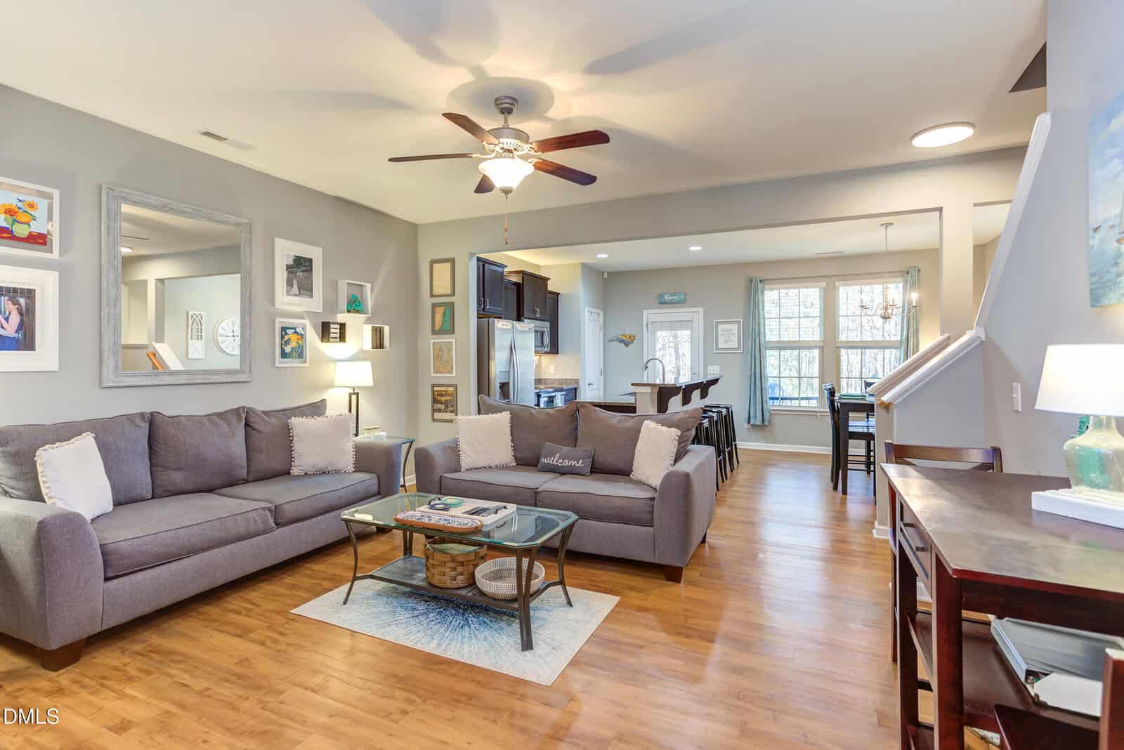 A cozy living room with two gray sofas, a glass coffee table, and wall art, seamlessly connected to a dining area and kitchen. The space features wood flooring, neutral colors, and natural light from large windows.