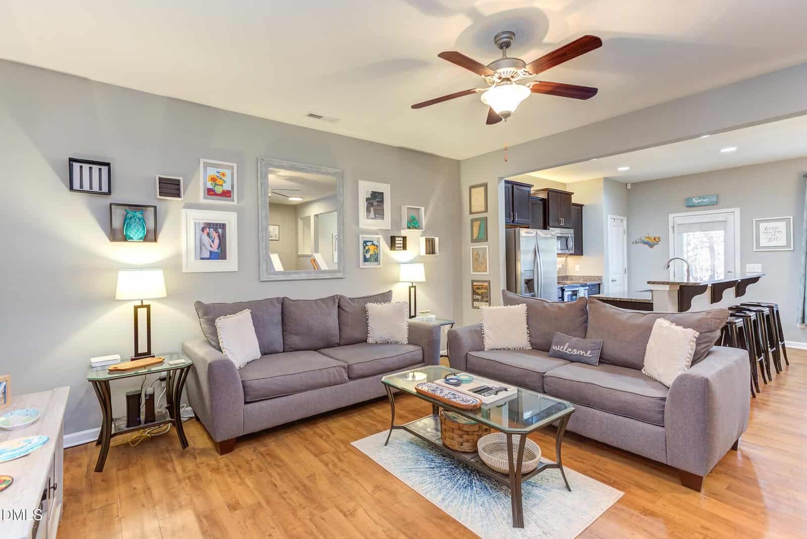 A cozy living room with two gray sofas, a glass coffee table, wall art, and decorative pillows. The space features wood floors, modern lighting, and an open view into a kitchen with bar stools and stainless steel appliances.