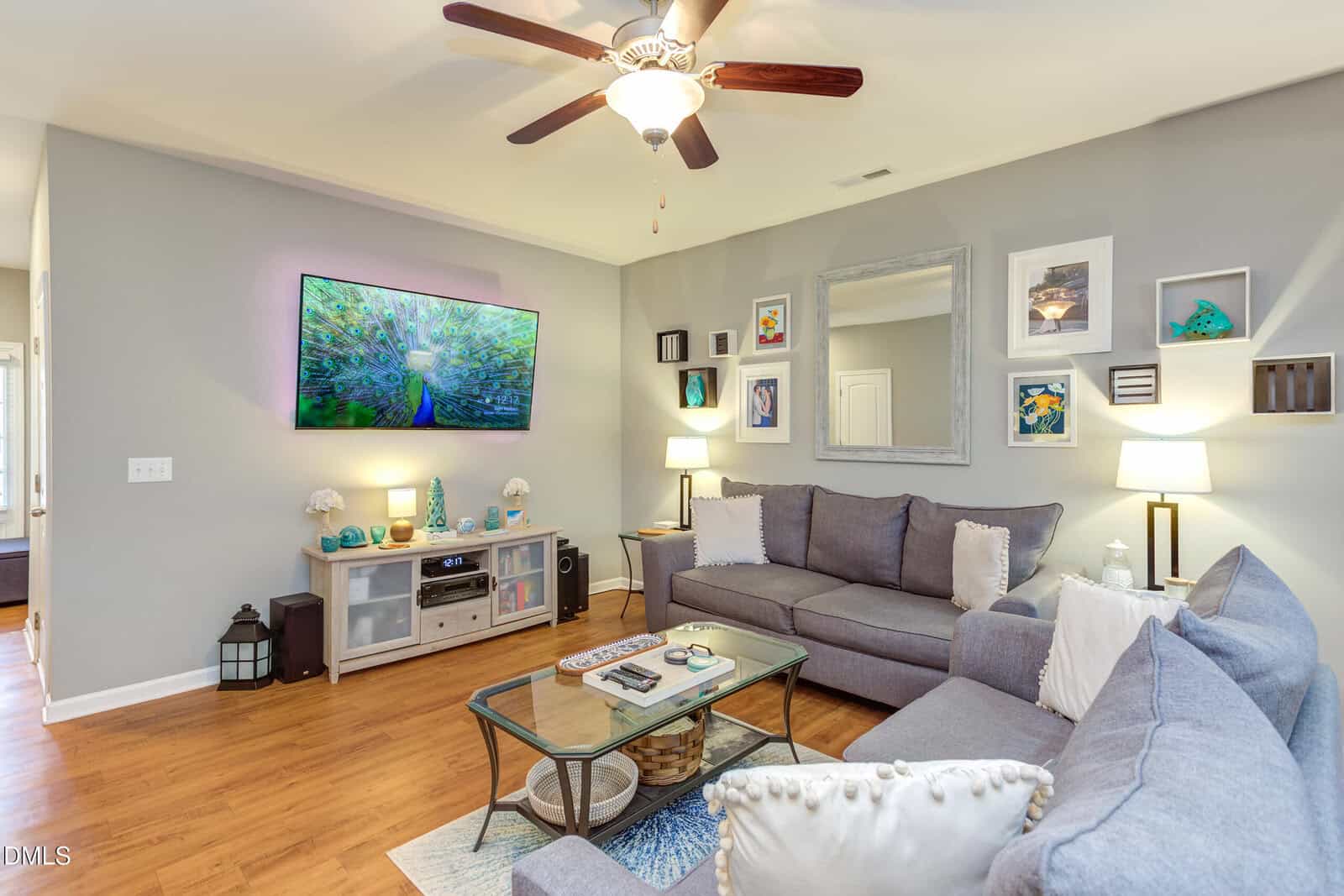 A cozy living room with gray sofas, a glass coffee table, wood floors, and a wall-mounted TV displaying scenic nature. The walls are decorated with framed art and shelves, and the room is softly lit with lamps.