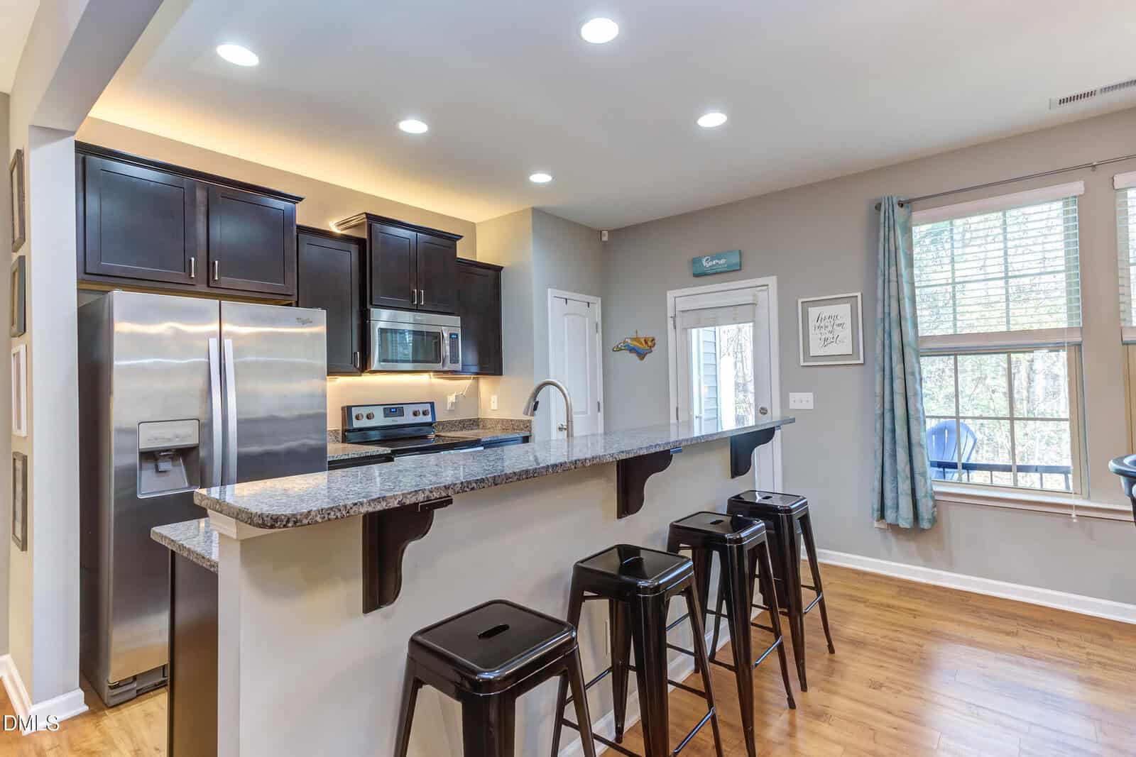 Modern kitchen with granite counter, stainless steel appliances, dark cabinets, and four black barstools. Large windows bring in natural light; wood flooring and light gray walls create a bright, welcoming space.