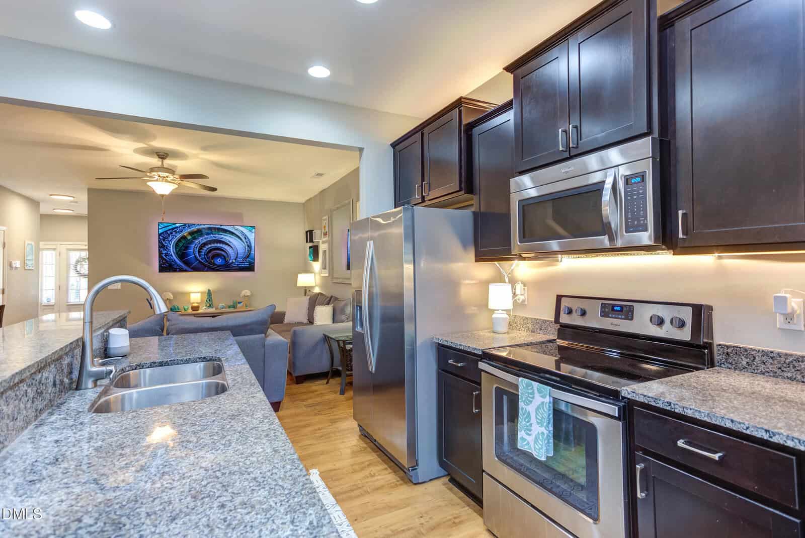 Modern kitchen with stainless steel appliances, dark cabinets, and granite countertops. The kitchen opens to a living room with a gray sectional sofa, TV, and ceiling fan. Bright lighting throughout.