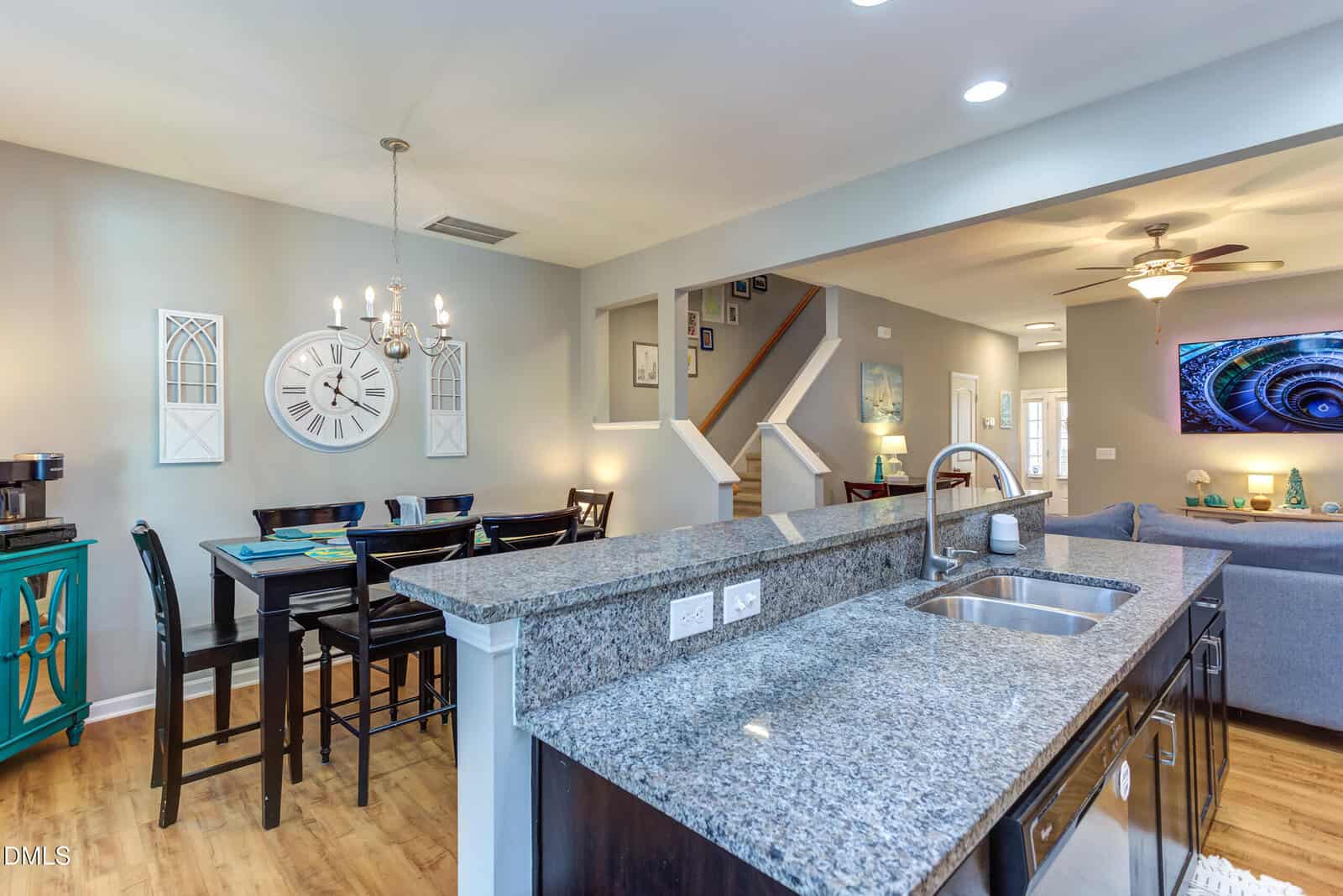 Open-concept kitchen with granite countertops overlooks a dining area with a large wall clock, dark dining set, teal side table, and a living room with gray sofa. Stairs lead up in the background. Warm, inviting lighting.