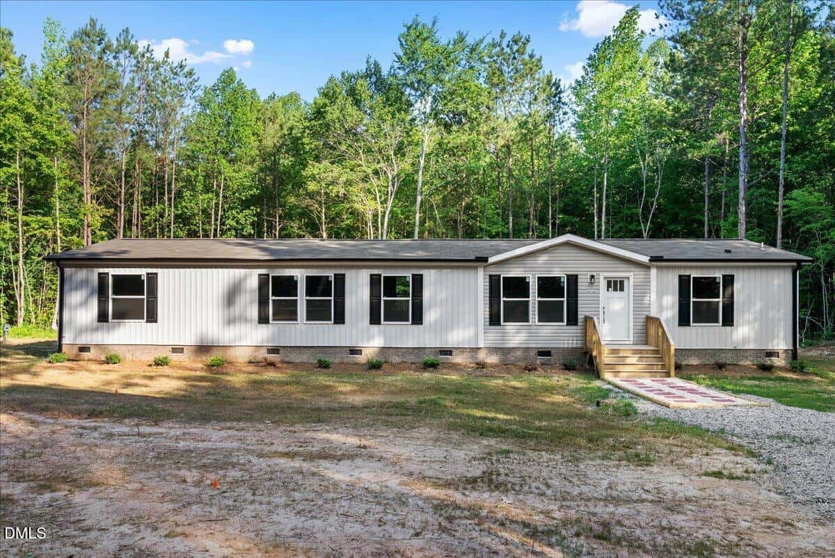 A long, single-story gray manufactured home with multiple windows and a small front porch sits in front of a wooded area and clear blue sky at 3712 N Carolina 58, Warrenton.