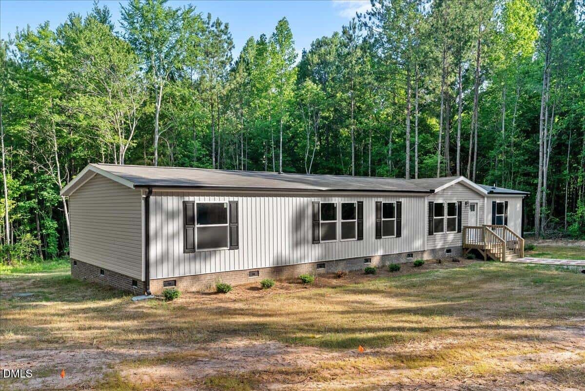 A beige manufactured home with dark trim sits on a cleared lot surrounded by tall trees and greenery, featuring multiple windows and a small front porch with steps.
