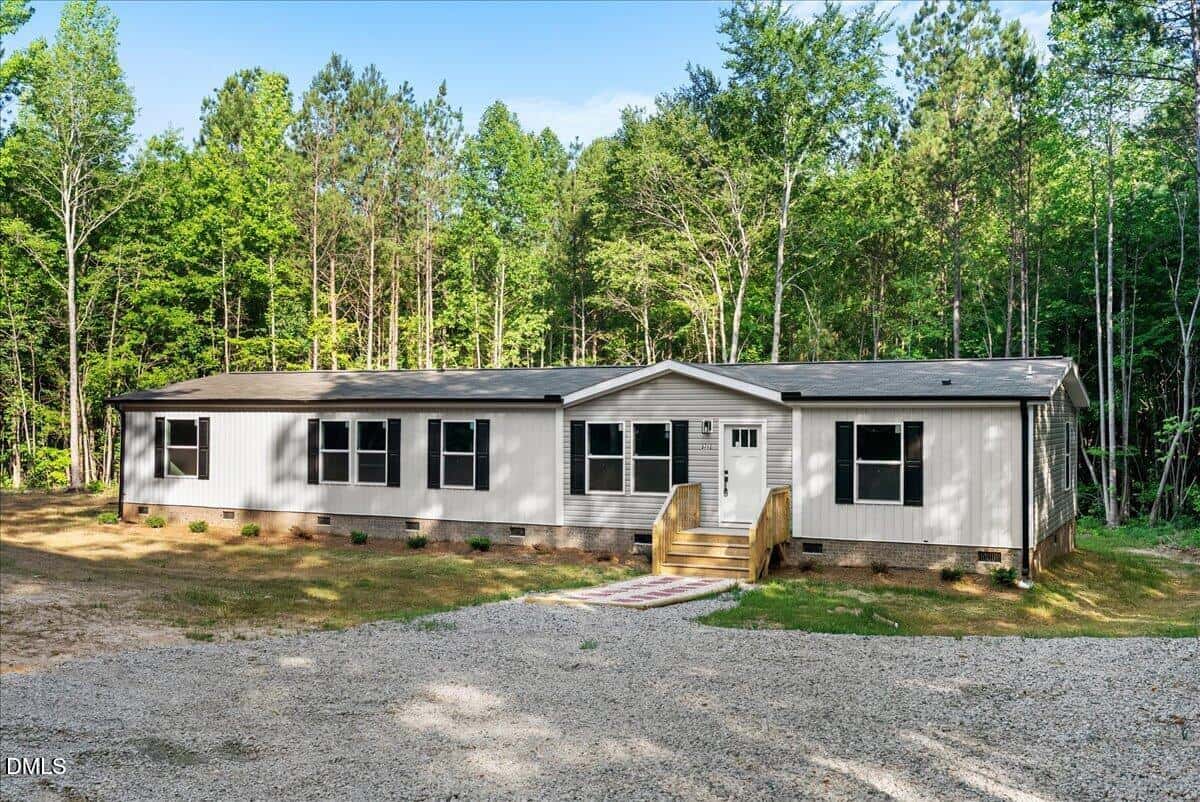 A white manufactured home with multiple windows sits in a clearing surrounded by tall trees. A small front porch with steps leads to the entrance, and a gravel driveway is in the foreground.