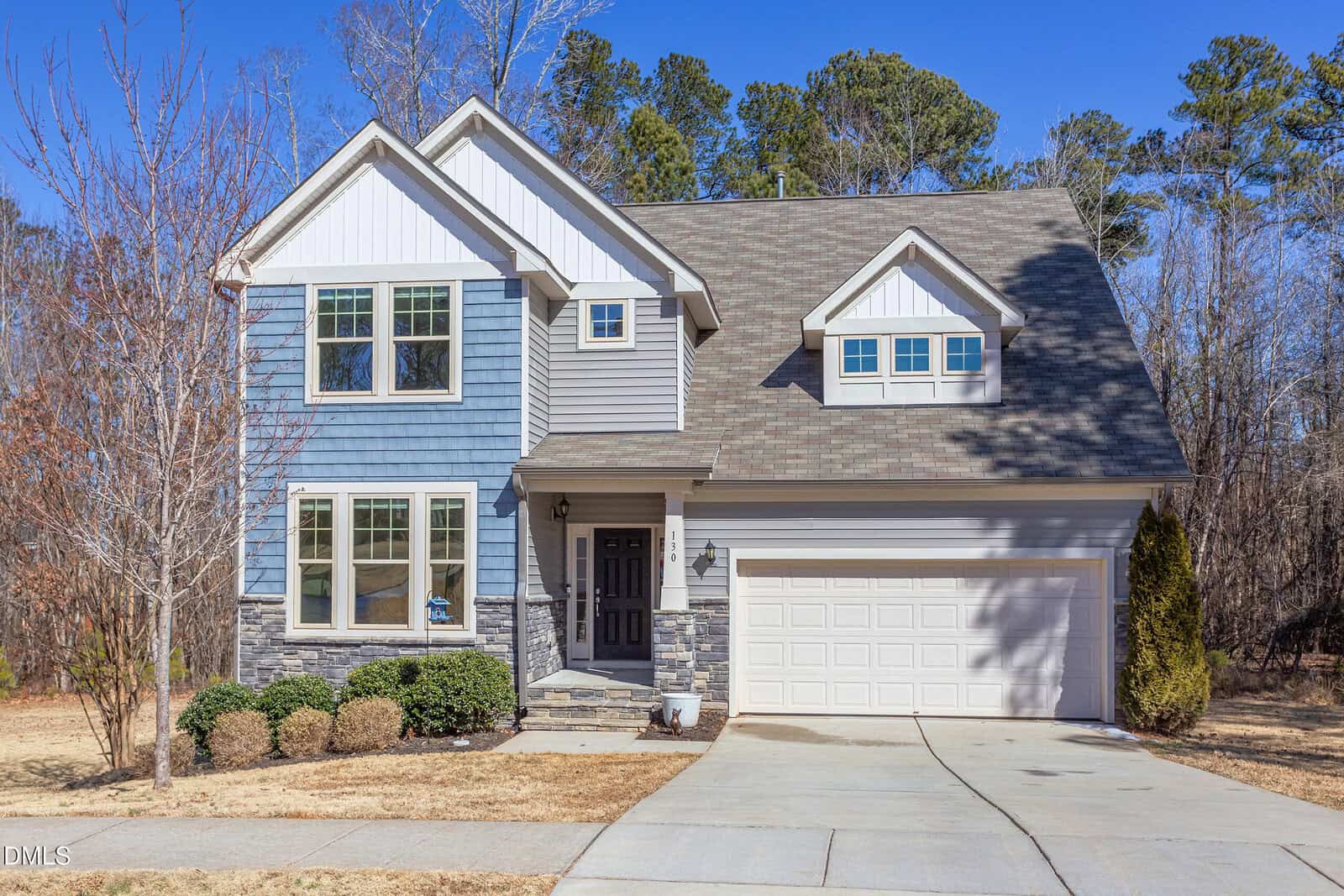Two-story suburban house with blue and gray siding, a gabled roof, double garage, and front porch situated in Franklinton at 130 N Ridge View Way. Leafless trees and shrubbery surround the yard, and a concrete driveway leads to the street. The sky is clear and blue.