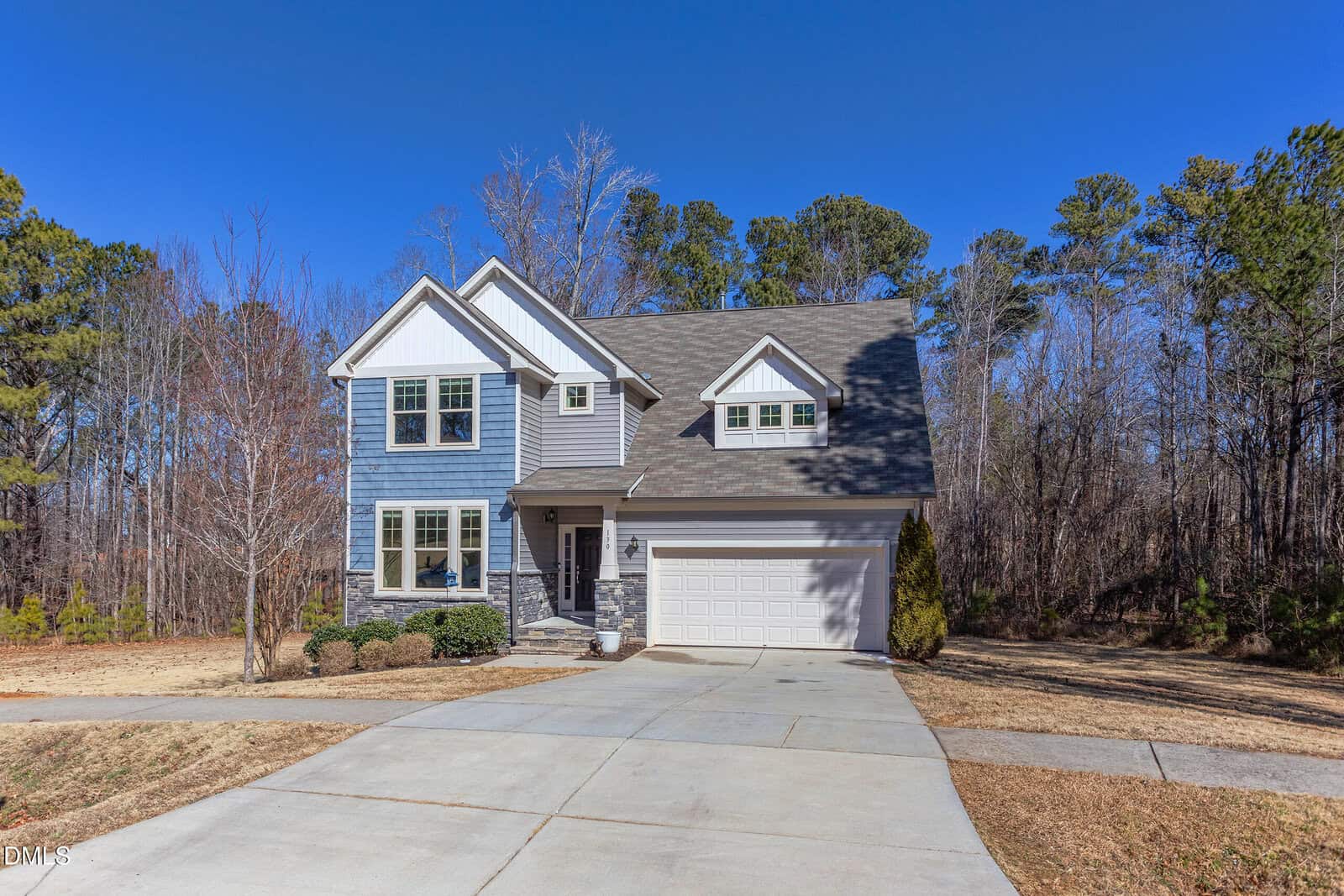 Two-story blue house with white trim, attached two-car garage, and stone accents. The house sits on a concrete driveway with a lawn in front and wooded area in the background under a clear blue sky.