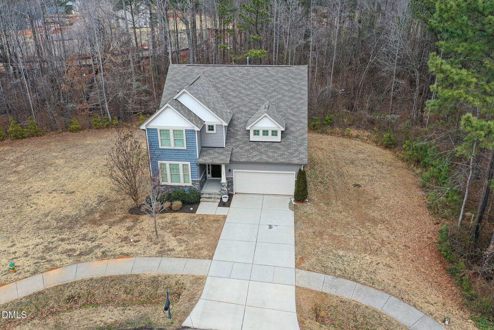 A two-story house with gray siding and a front porch sits at the end of a curved driveway. The surrounding yard is mostly brown grass and bordered by tall trees in the background.