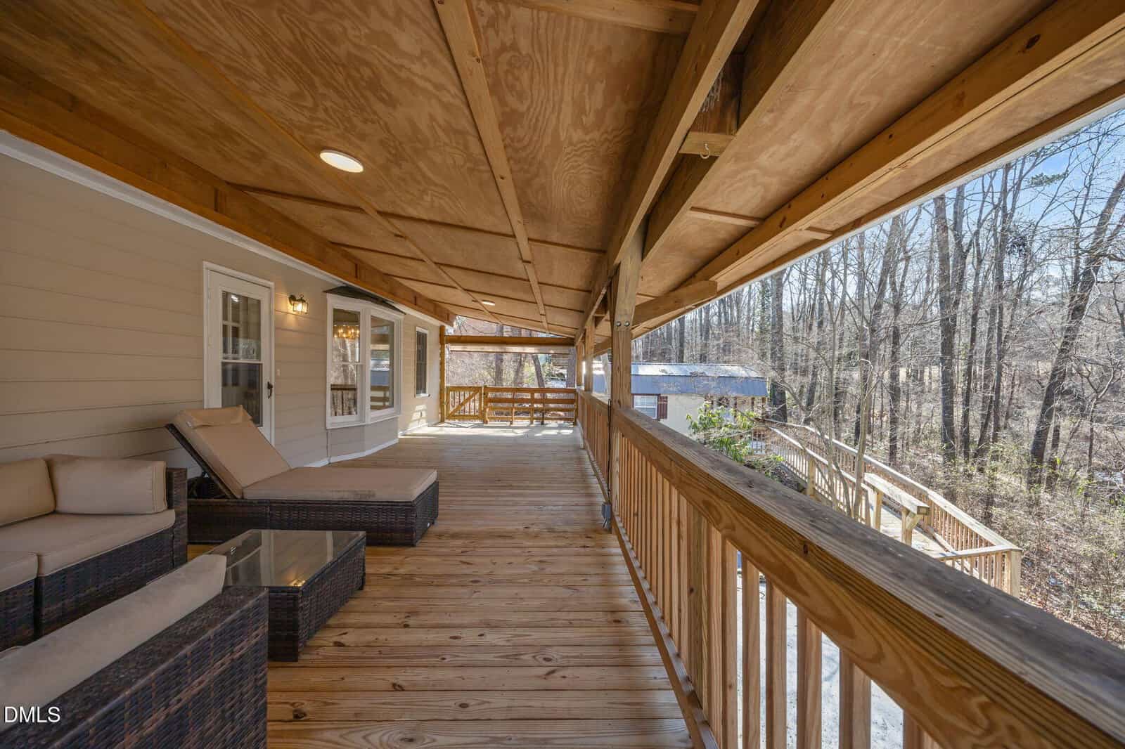 A spacious covered wooden deck with wicker lounge chairs, glass-top tables, and railings overlooks a wooded area on a sunny day. The ceiling has exposed beams and recessed lighting.