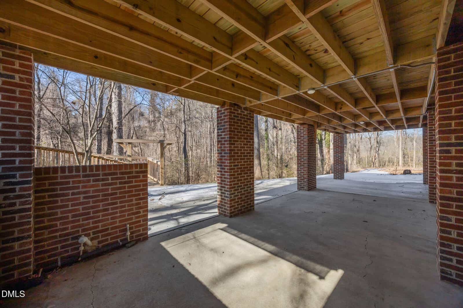 View of a covered patio with a wooden ceiling, exposed beams, brick columns, and concrete floor, overlooking a backyard with leafless trees and a wooden structure in the distance.