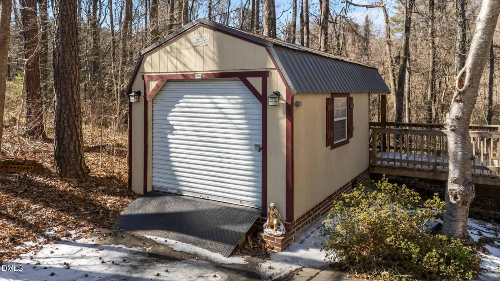 A small beige shed with a brown roof and trim, featuring a roll-up garage door and ramp, sits in a wooded area with snow and fallen leaves on the ground. A wooden deck is visible to the right.