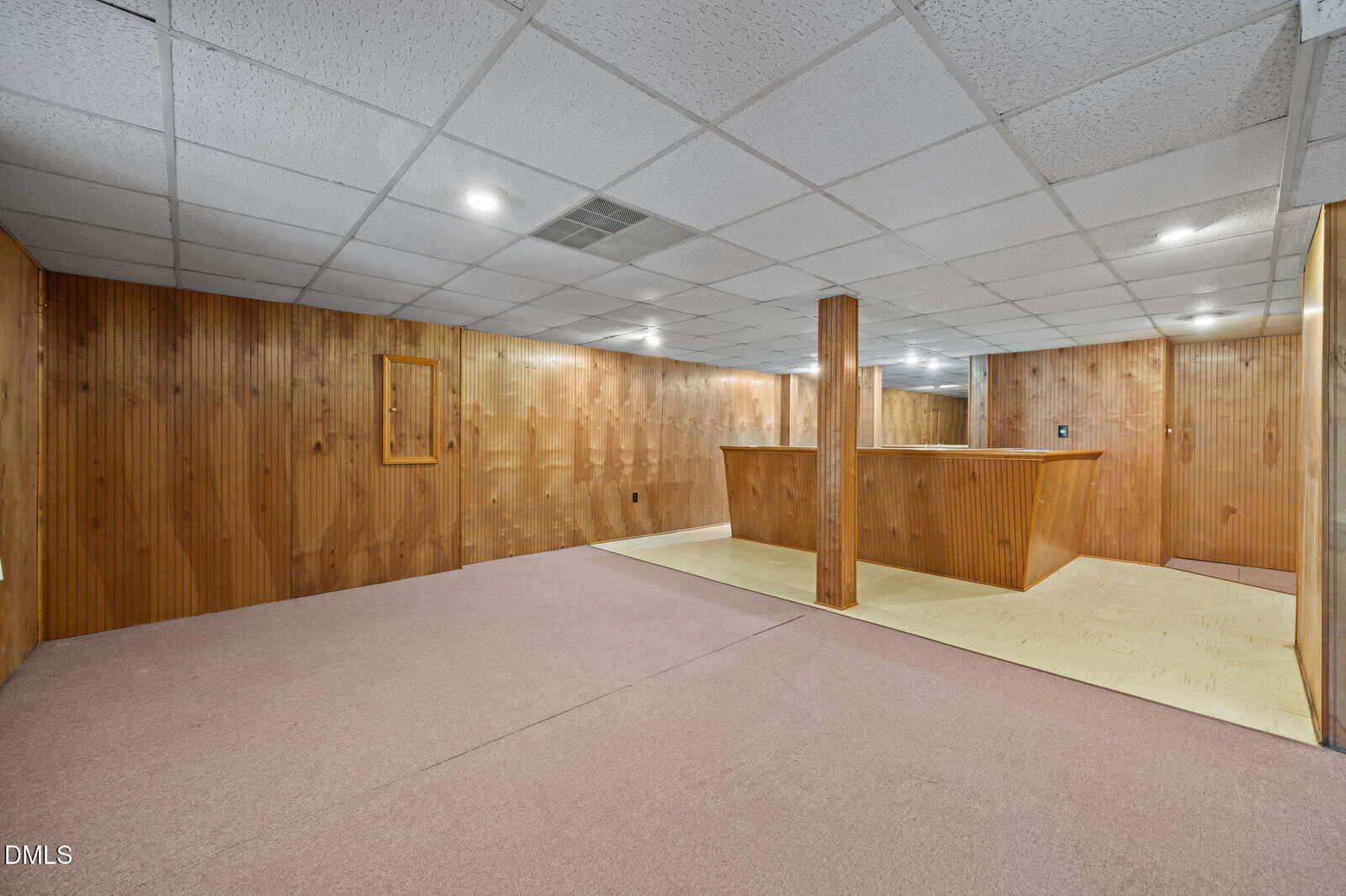 Basement with wood-paneled walls, drop ceiling tiles, recessed lighting, a support column, and a half-wall separating a carpeted area from a linoleum floor section.