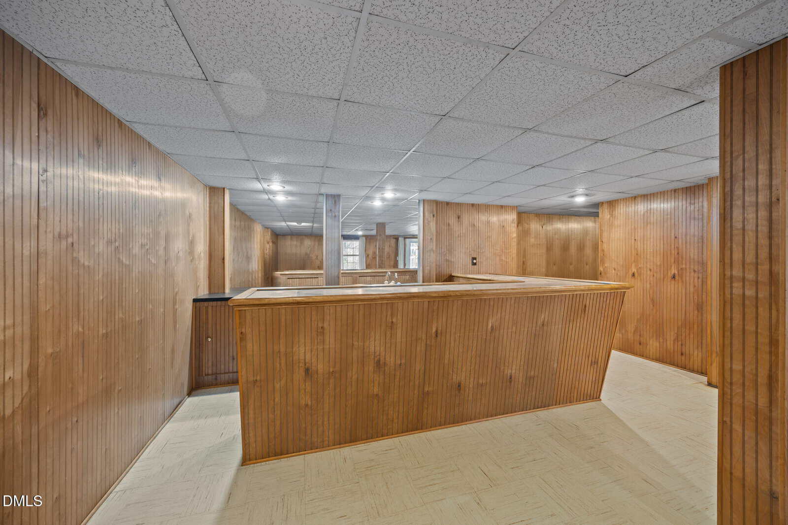 Basement room with wood-paneled walls, drop ceiling with recessed lights, and a large wooden counter or bar area. Reflective mirror panels are visible on the back wall. Light-colored linoleum flooring.