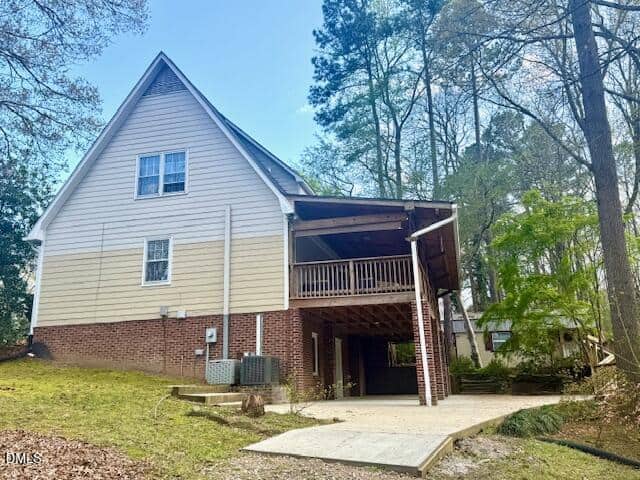 A two-story house with beige siding and a brick foundation features a covered carport and an upper-level balcony. Surrounded by trees, the home sits on a sloped yard with a concrete driveway and steps leading to the entrance.