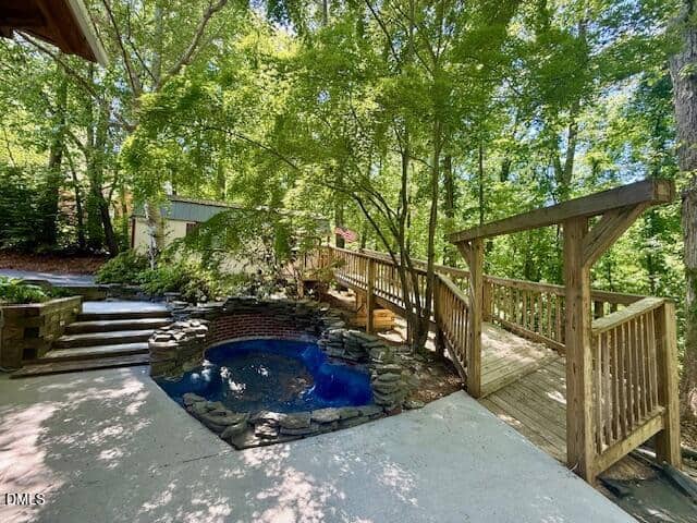 A small stone-lined pond with blue water sits next to a wooden ramp and steps surrounded by lush green trees and dappled sunlight in a peaceful backyard setting.