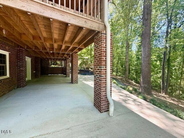 Covered patio area with brick columns and exposed wooden beams underneath a deck, next to a concrete driveway and surrounded by tall trees and greenery.