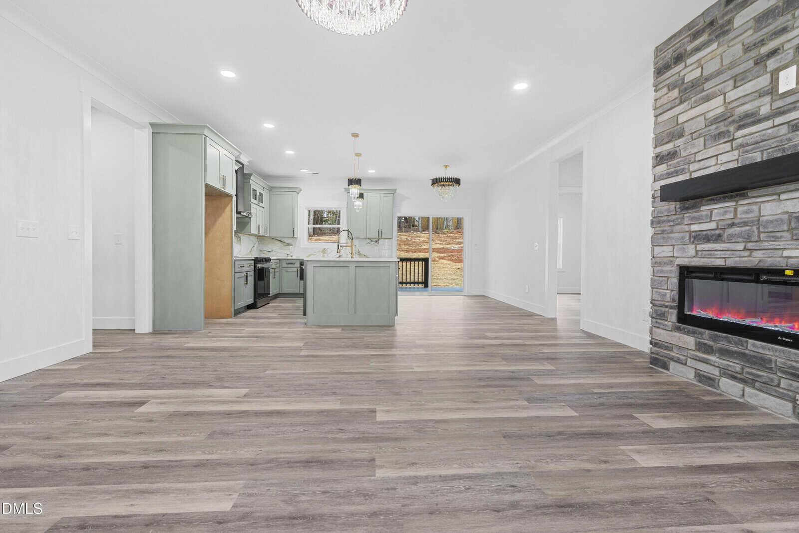 Open-concept kitchen and living area with wood floors, a stone fireplace with electric insert, modern light fixtures, and large windows overlooking a deck and yard. Light gray cabinets and an island are featured in the kitchen.