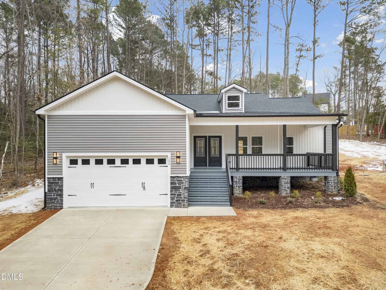 Single-story house with gray siding, a front porch, white garage door, stone accents, and front steps. Surrounded by dry grass, sparse snow, and tall trees in the background under a partly cloudy sky.
