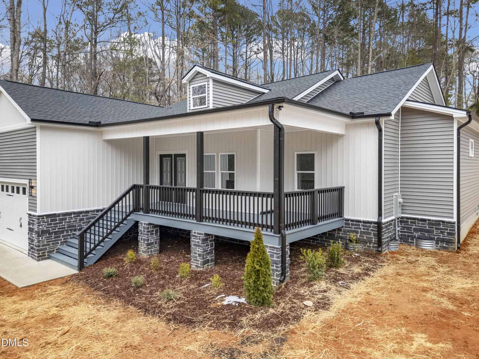 Single-story house with gray siding, black trim, covered front porch, black railings, and stone pillars. Mulched garden bed and young plants in front, surrounded by trees.