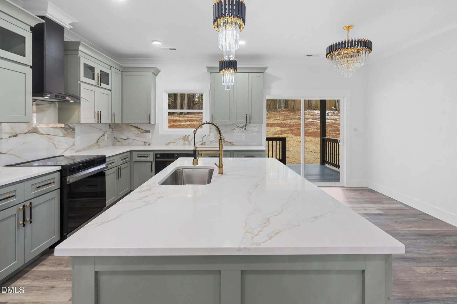 Modern kitchen with gray cabinets, marble countertops and backsplash, gold hardware, a large island with sink, two pendant lights, and sliding glass doors opening to a yard with brown grass.