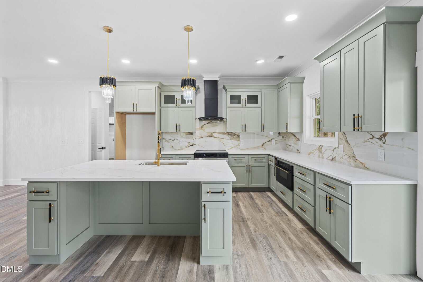 Modern kitchen with sage green cabinets, marble countertops and backsplash, gold fixtures, pendant lights, and wood-look flooring. Large island features a built-in sink and ample storage, with stainless steel appliances throughout.